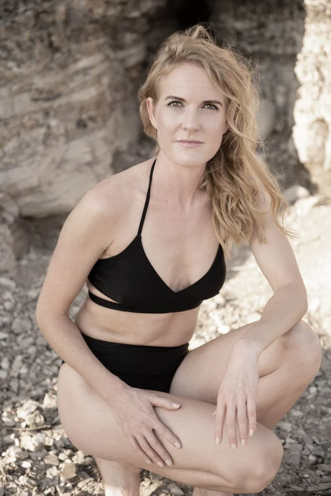 A woman with blonde, wavy hair posing outdoors on rocky terrain, wearing a black bikini.