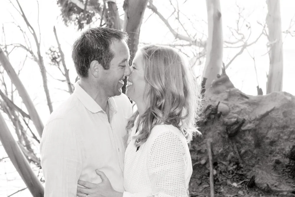 A couple sharing a kiss outdoors, with trees and a large rock in the background.