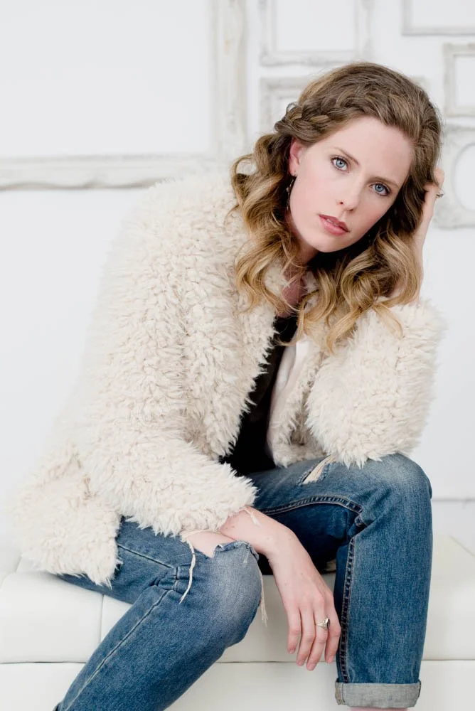 A woman with wavy blonde hair and blue eyes sitting on a white sofa, wearing a cream-colored fuzzy coat and ripped jeans, in a bright room with white decor.