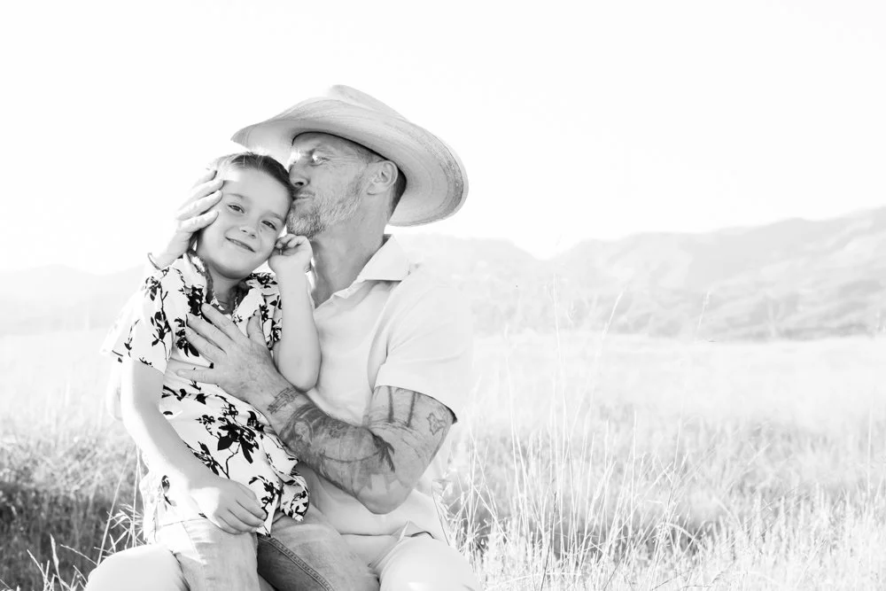 A man and young girl share a tender moment outdoors on a grassy field, with the man kissing the girl on her cheek.