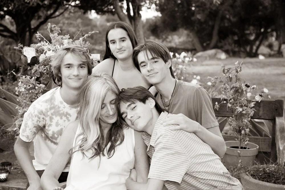 A group of six people, five children and one adult woman, gathered outdoors near plants and trees.