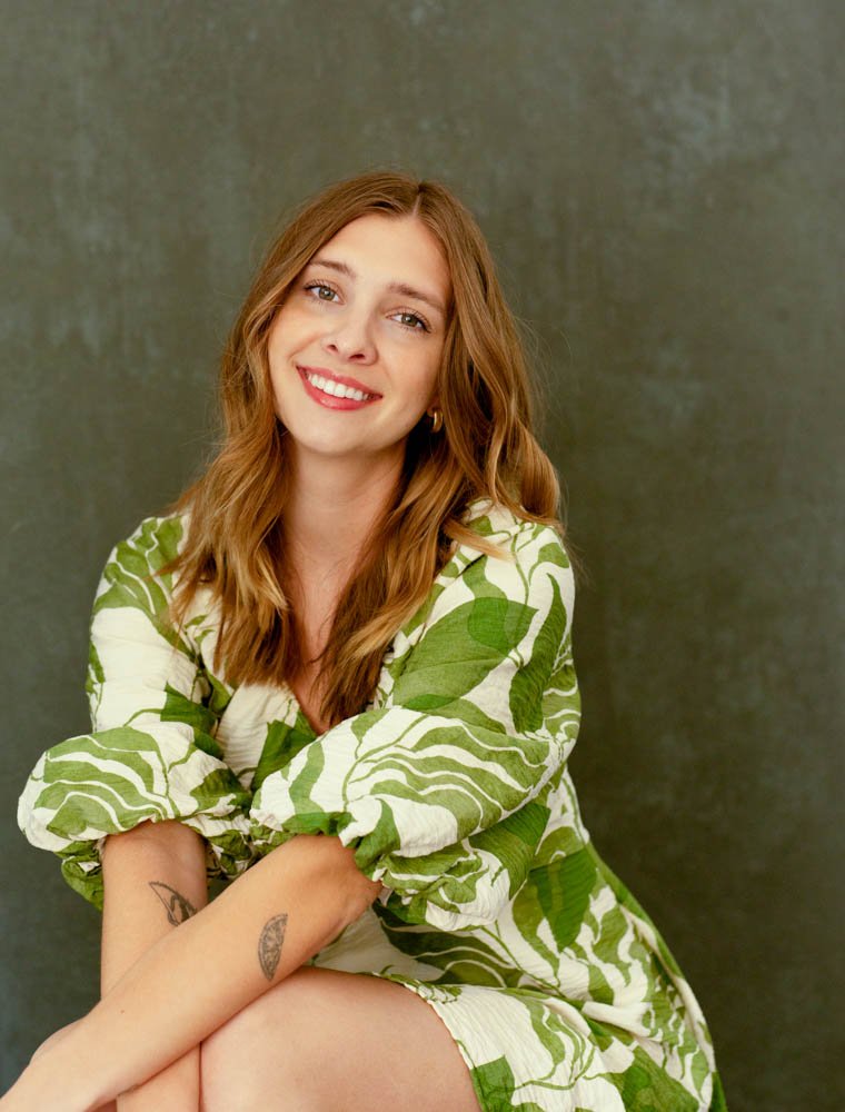 A young woman with long red hair, smiling, wearing a green and white leaf-patterned dress, sitting against a gray background.