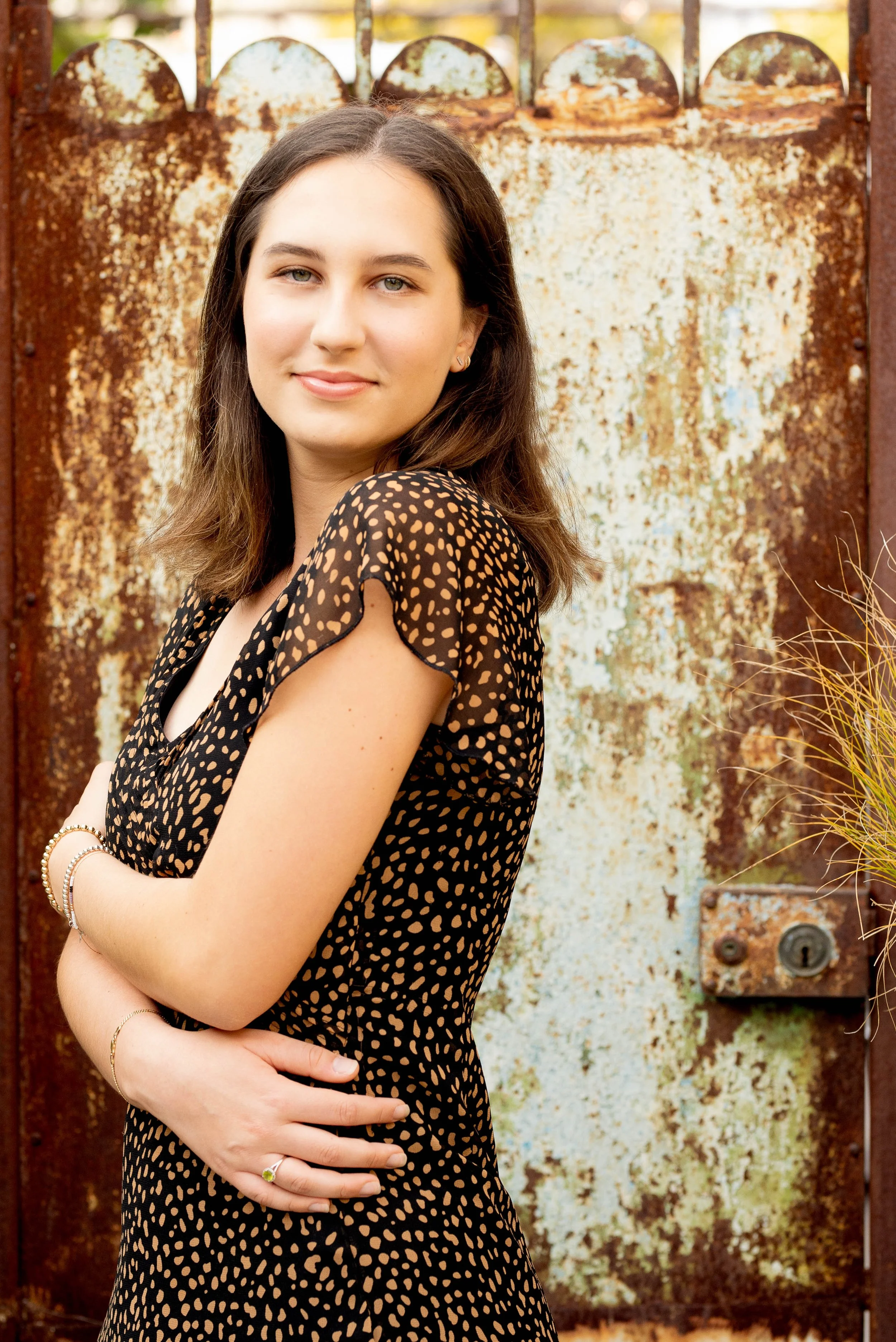 A young woman with shoulder-length brown hair and light skin, smiling softly with arms crossed, standing in front of a rusted metal gate.