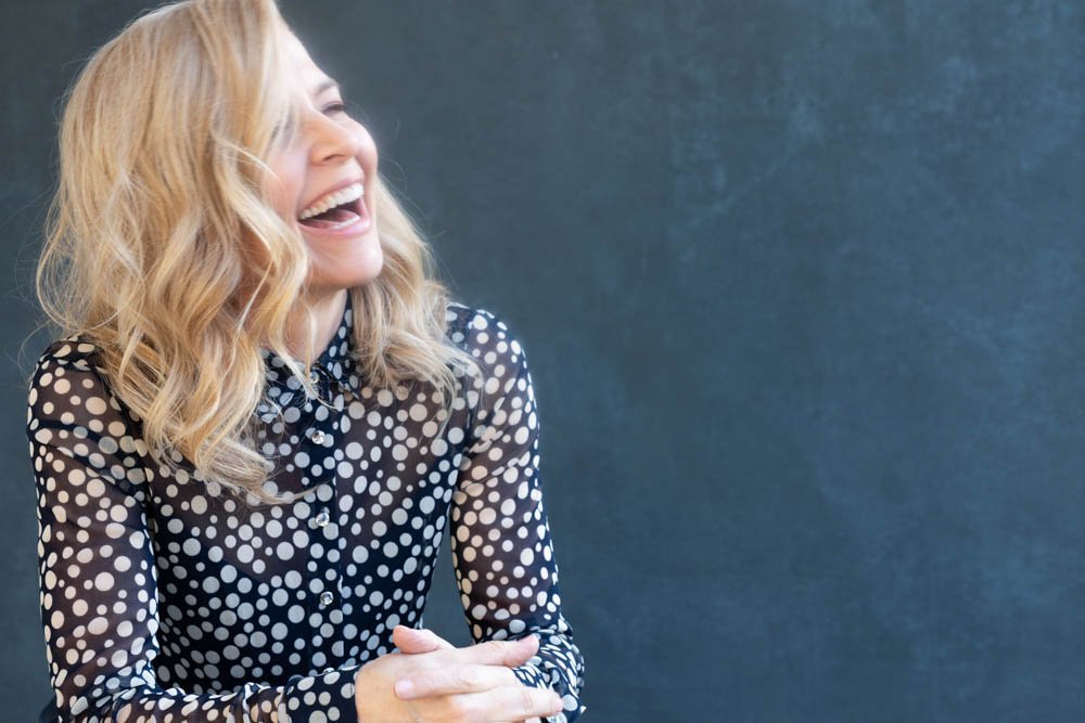 A woman with blonde, wavy hair smiling and laughing, wearing a black blouse with white polka dots, sitting against a dark background.