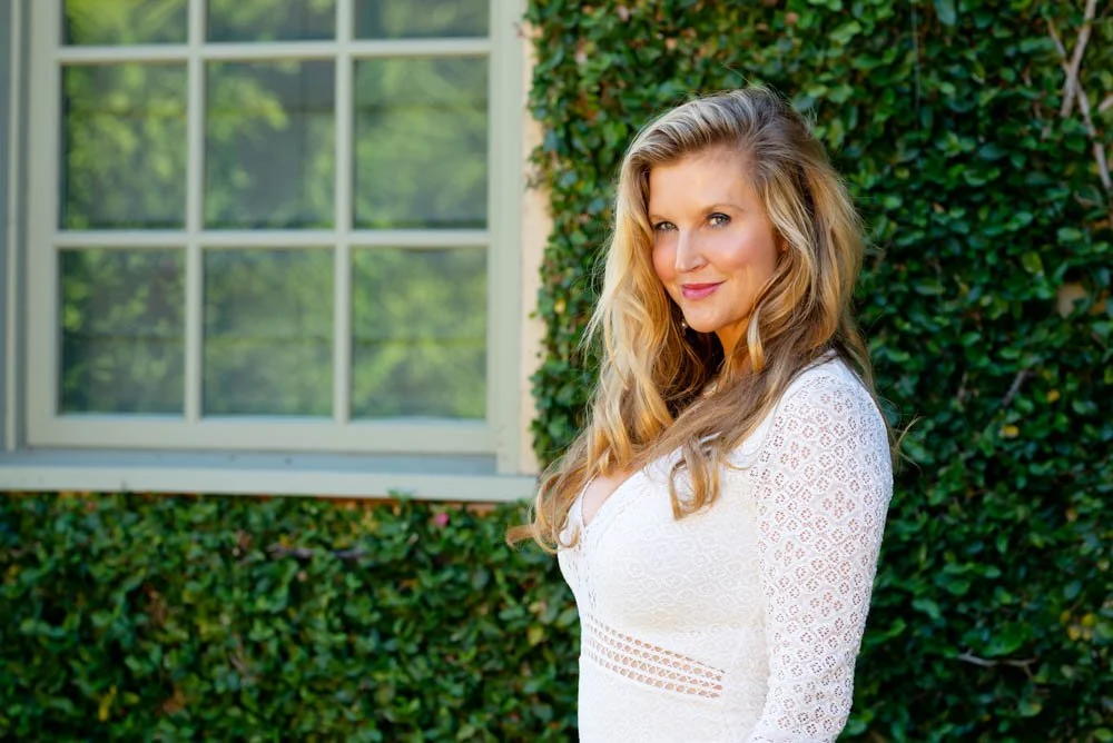 A woman with long, wavy blonde hair standing outdoors near a window and green hedge, wearing a white lace dress, smiling softly.