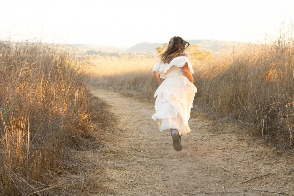 A young girl in a white dress running down a dirt path through dry grass.