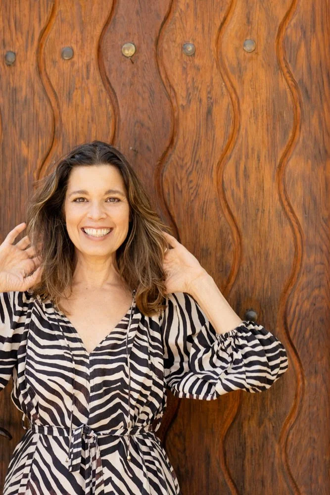 A woman smiling and posing with her hands near her ears in front of a textured wooden background.