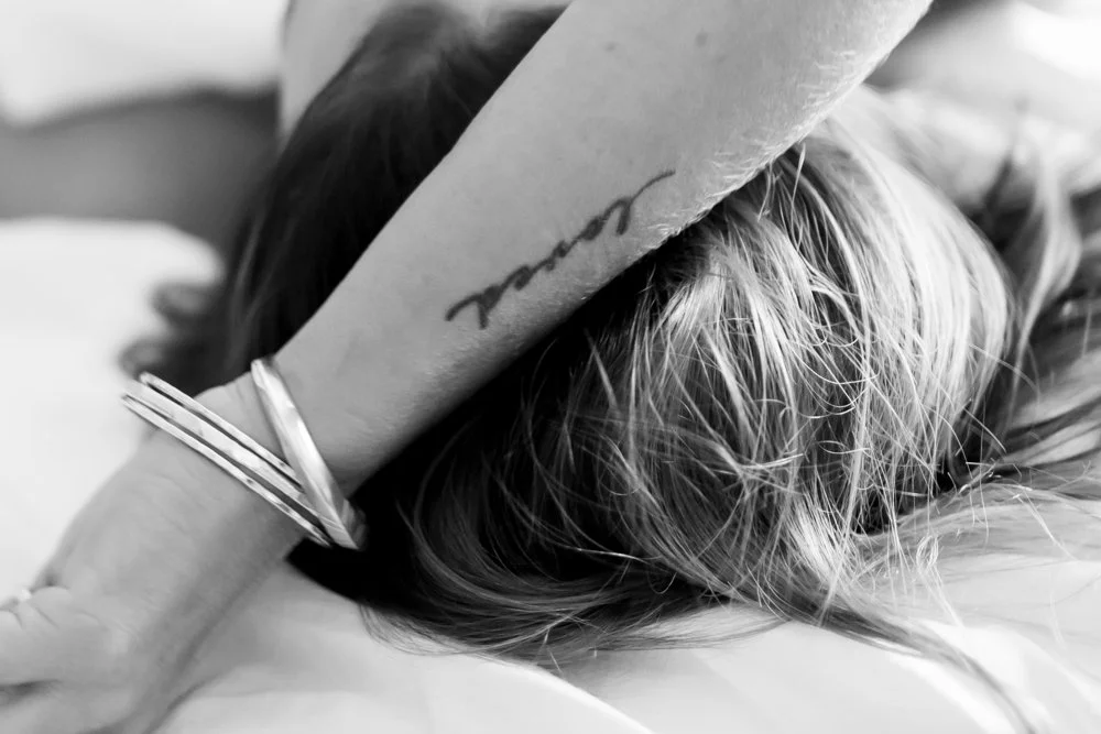 A person with tattoos and silver bangles resting their arm on a person's head, who has long hair and is lying down.   Boudior.   Sensual photo.