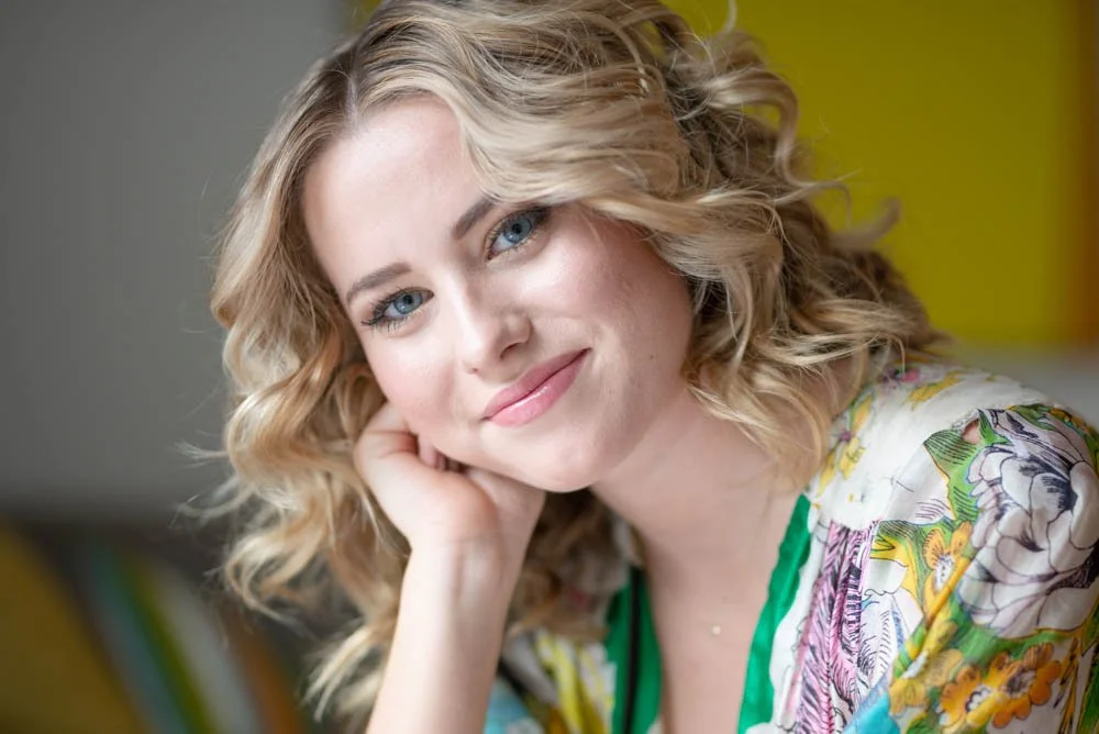 Close-up of a woman with curly blonde hair smiling softly and resting her head on her hand, wearing a colorful floral top.
