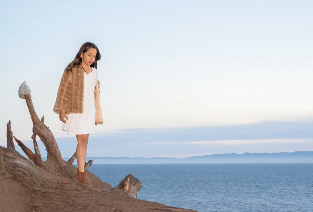 A girl standing on a piece of driftwood on a beach, looking down. She is wearing a white dress, a brown furry jacket, and brown shoes, with the ocean and distant mountains in the background.