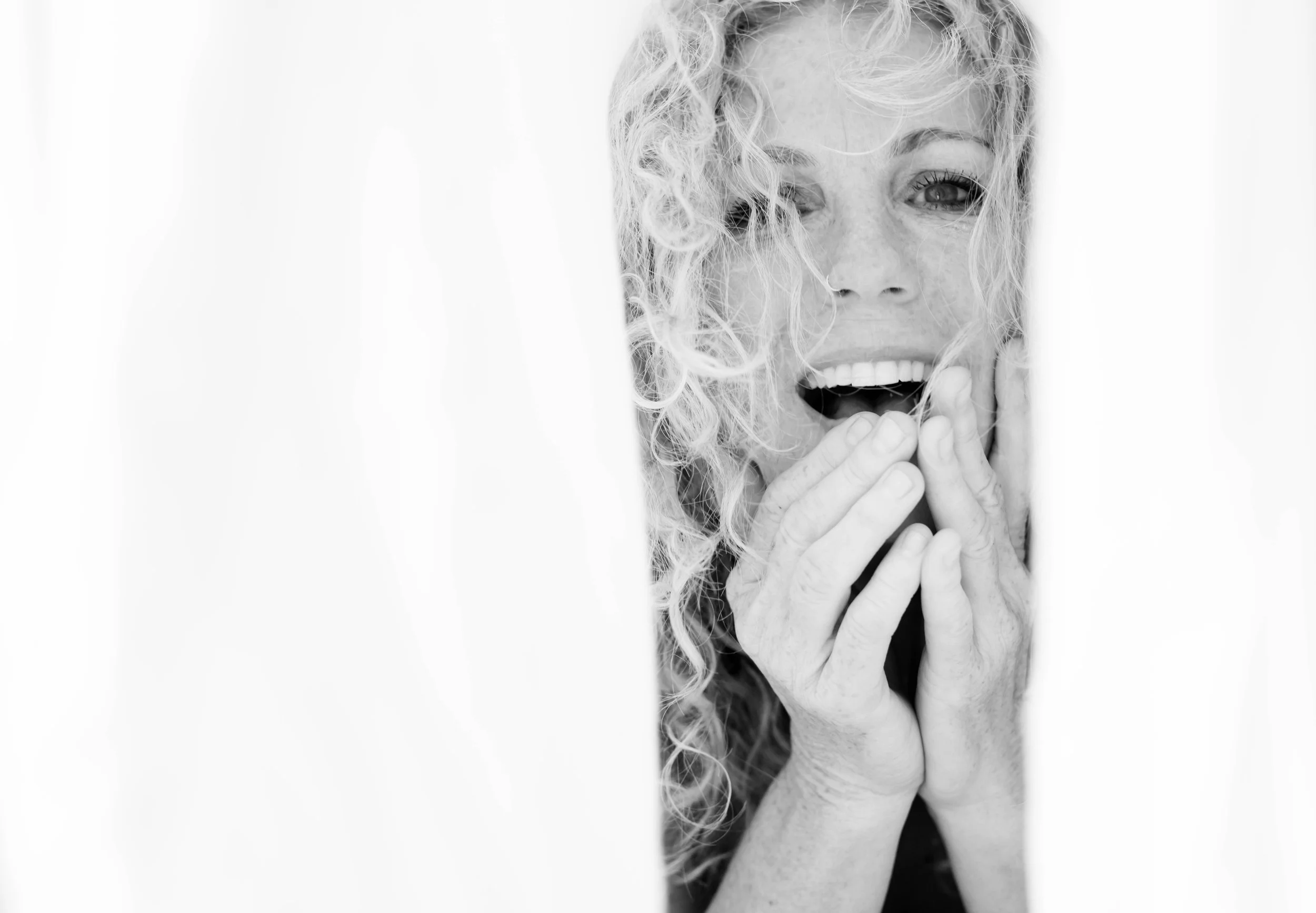 A woman with curly hair peeking through a curtain, smiling with her hands near her face.