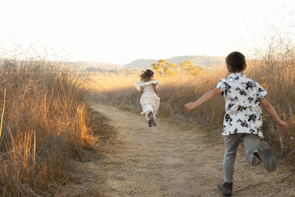 Two children, a girl and a boy, running on a dirt trail through a field of tall dry grass during sunset or late afternoon.