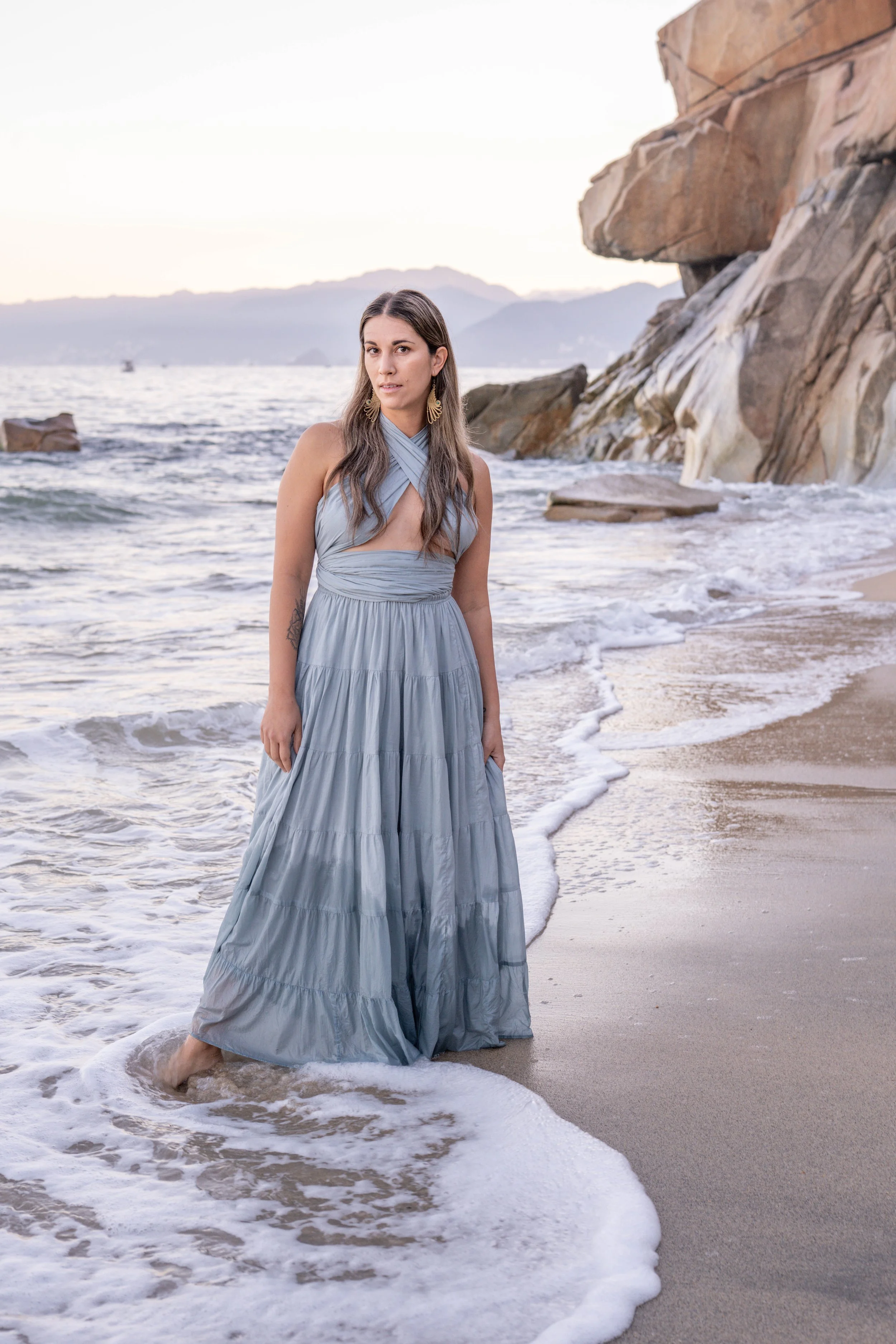 A woman in a long, light blue dress standing in shallow ocean water on a sandy beach near large rocks and cliffs, with mountains and a boat in the background during sunset.