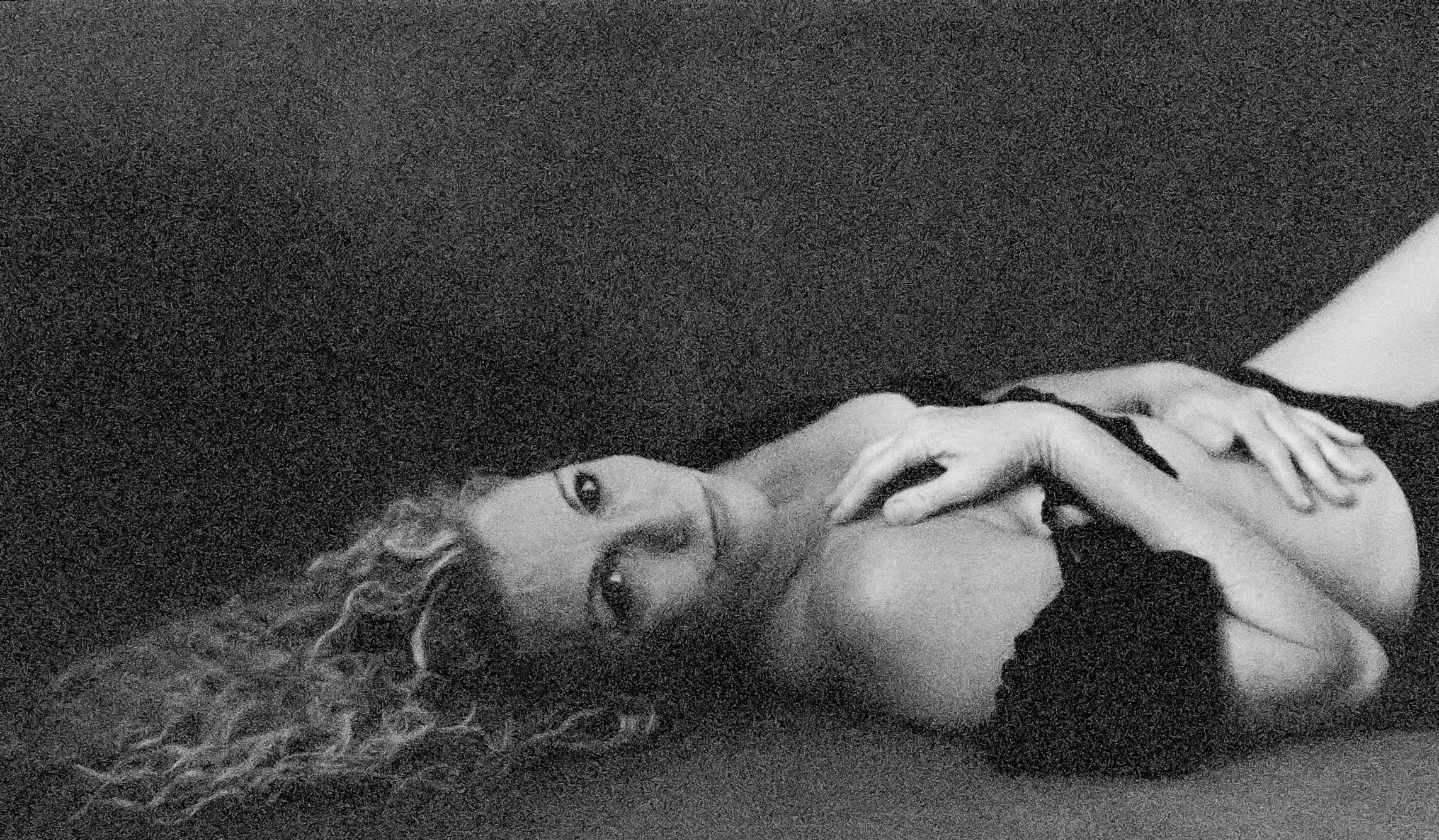 A woman lying on her side on a dark surface, with her head resting on the ground, looking at the camera. She has curly hair and is wearing a black top.