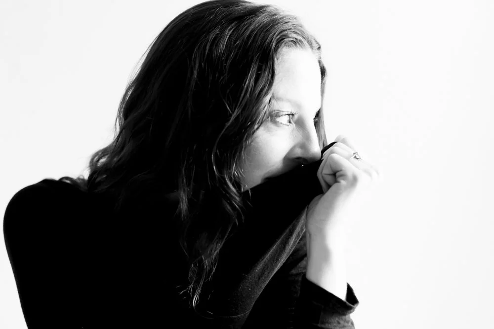 Black and white portrait of a woman with wavy hair, holding her sweater up to her face, partially covering her mouth.