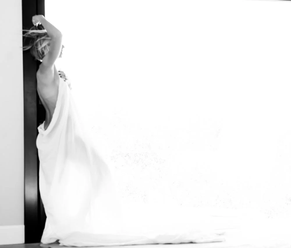A woman leaning against a wall with a white sheet draped over her, one arm raised above her head and her face turned away.