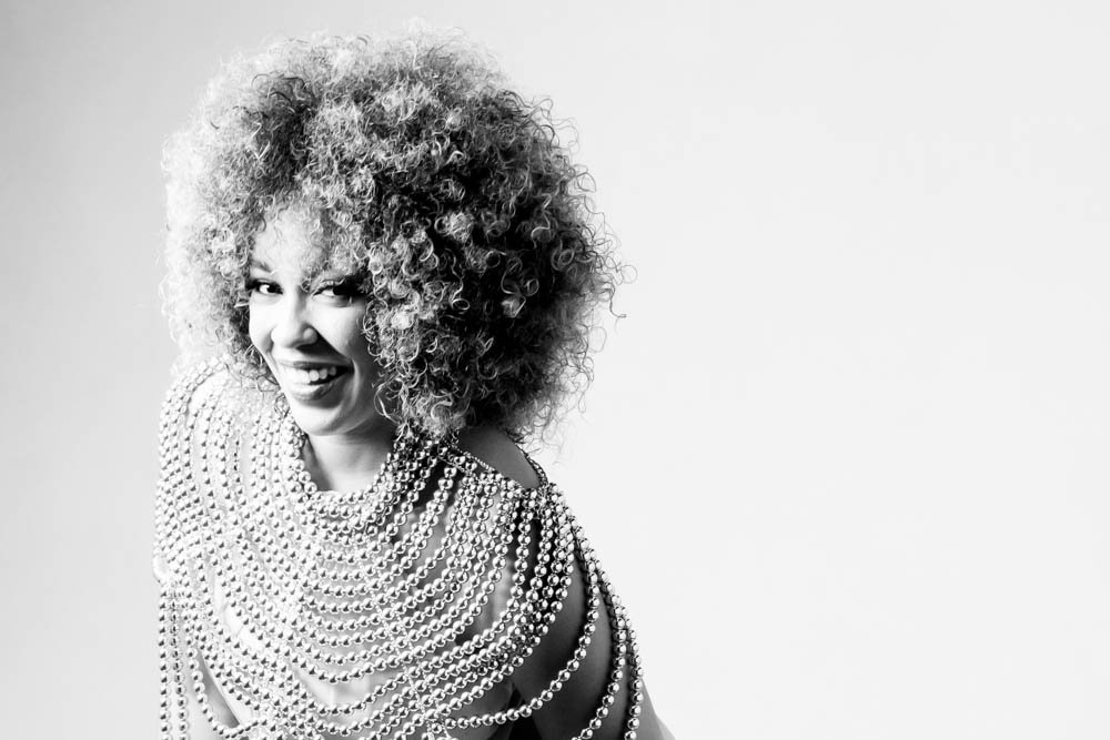 A woman with curly hair smiling while wearing layered pearl necklaces in a black-and-white photo.