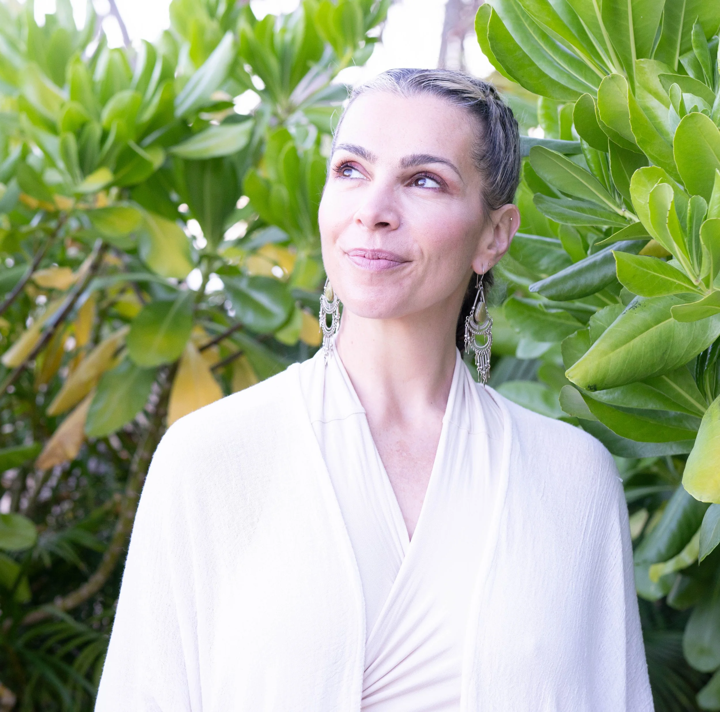 A woman with dark hair styled in a braid, wearing large silver earrings and a white top, standing among green leafy plants and looking off into the distance.