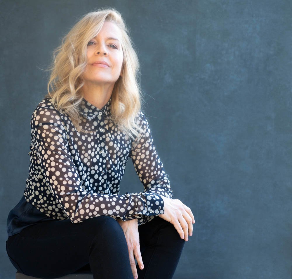 A woman with blonde, wavy hair sitting against a dark gray background, wearing a black blouse with white polka dots and black pants.