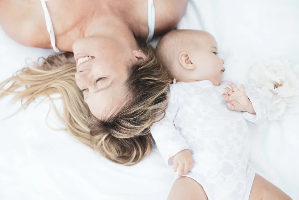 A smiling woman and a sleeping baby lying on a white bed.