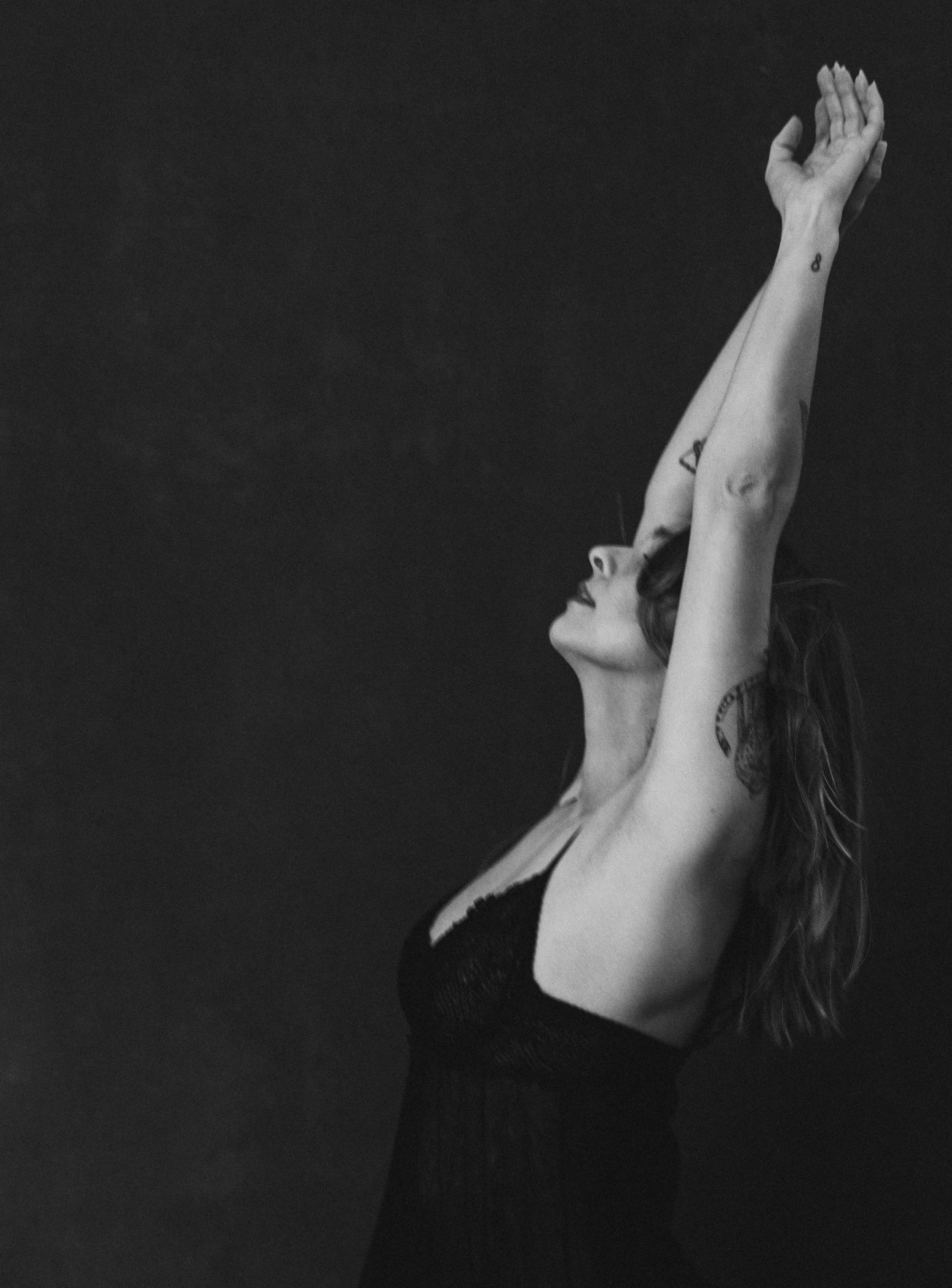 A woman with tattoos and wavy hair, wearing a black strapless dress, stands against a dark background with arms raised above her head and eyes closed.