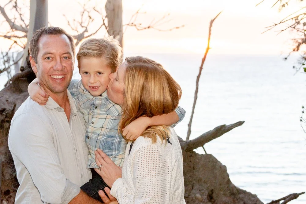 A family of three, a father, mother, and young son, at a beach during sunset, with trees and water in the background.