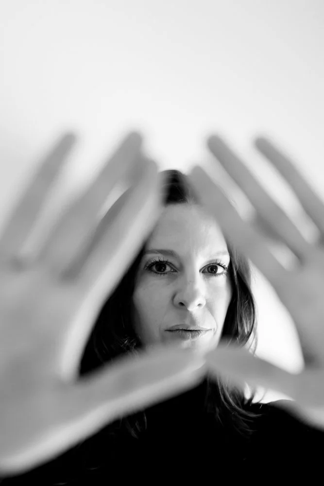 A black-and-white photo of a woman with dark hair, holding her hands in front of her face in a triangle shape, with her eyes looking through the opening.