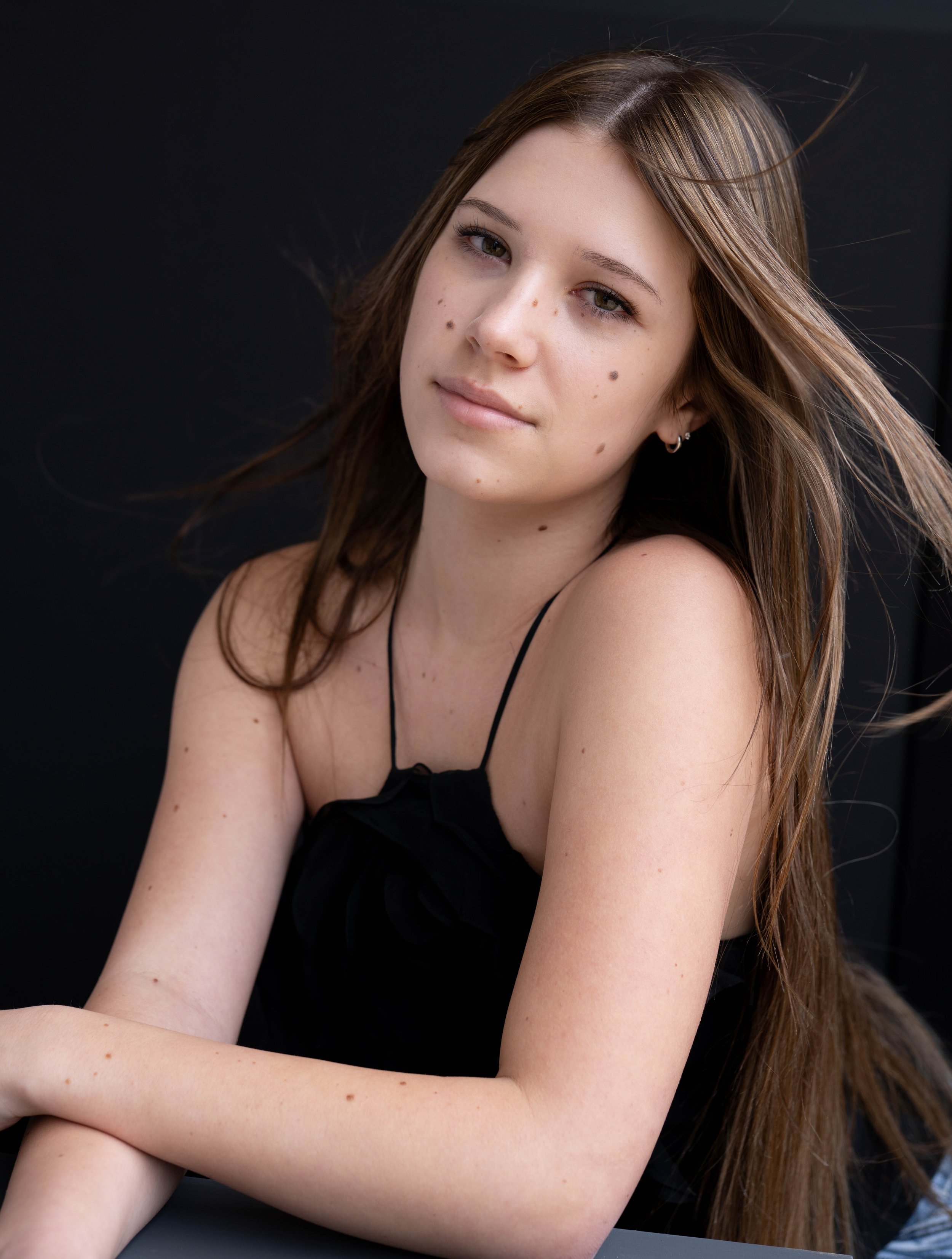 A young woman with long brown hair, wearing a black spaghetti-strap top, sitting against a black background.
