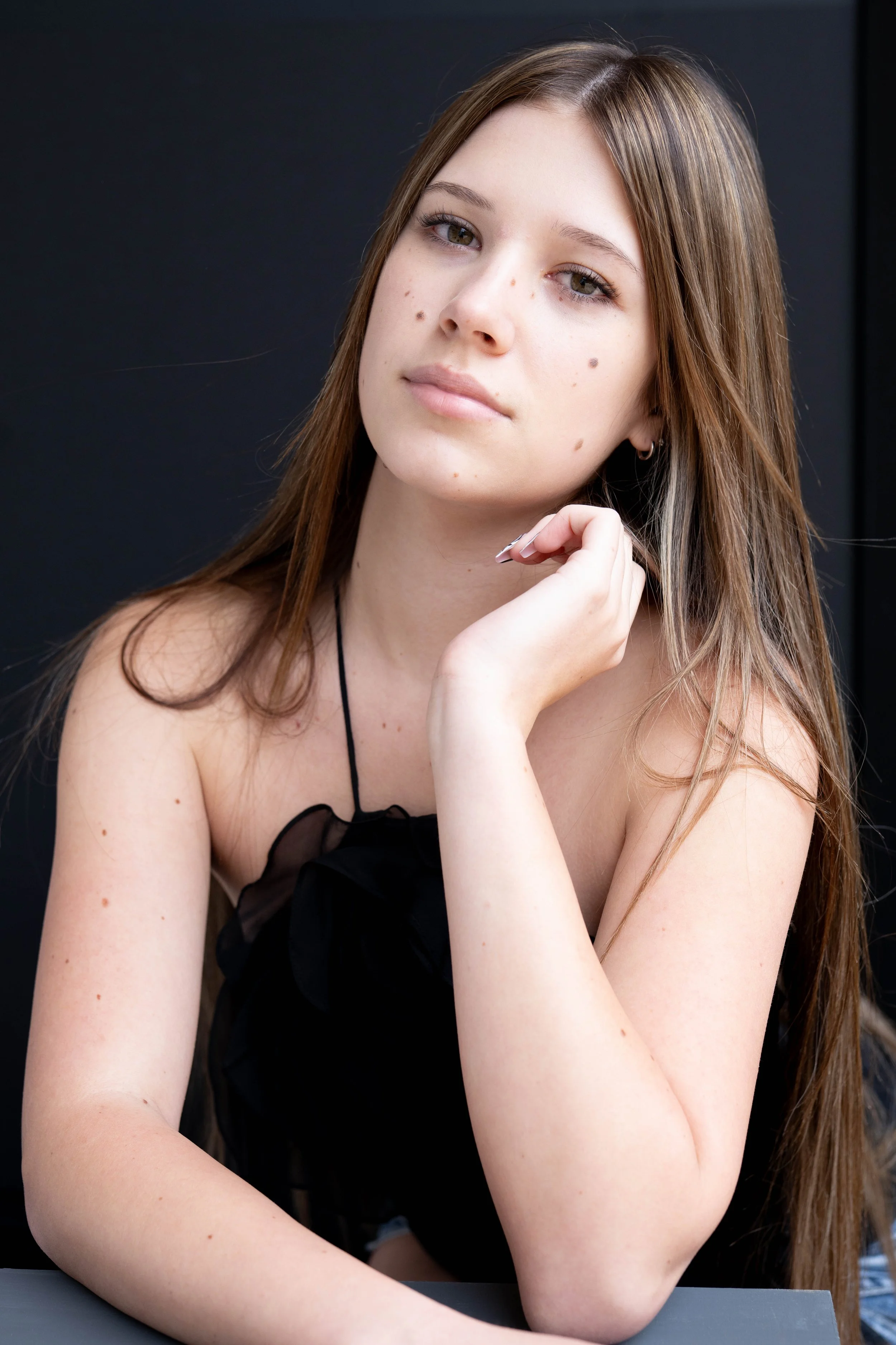 Young woman with long brown hair and multiple moles on face, wearing a black top, sitting at a table with a black background.