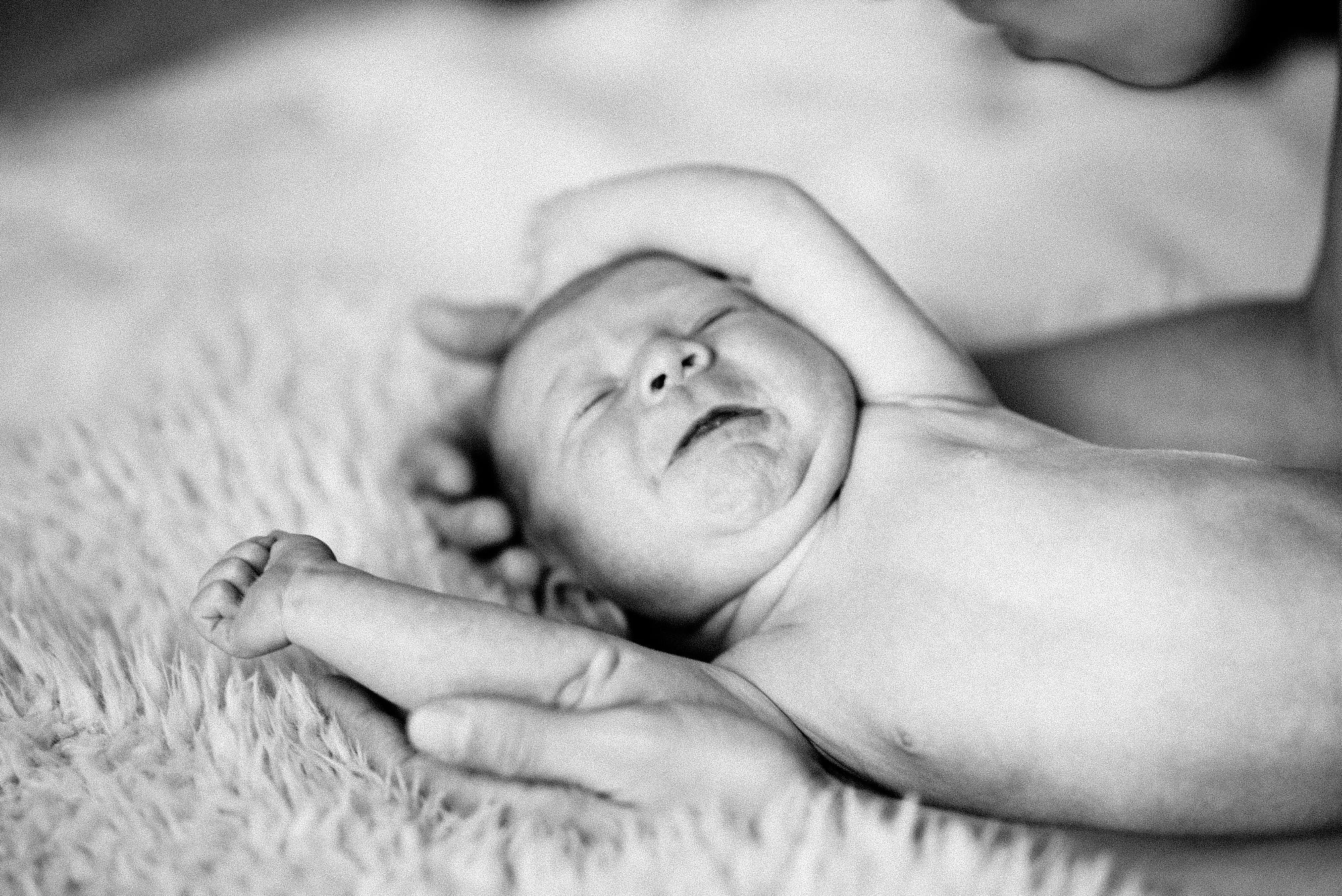 Black and white photograph of a newborn baby lying on a soft textured surface. The baby is holding an adult's hand, with the adult gently touching the baby's head. The baby has a peaceful, slightly open-mouthed expression, and the scene conveys tende