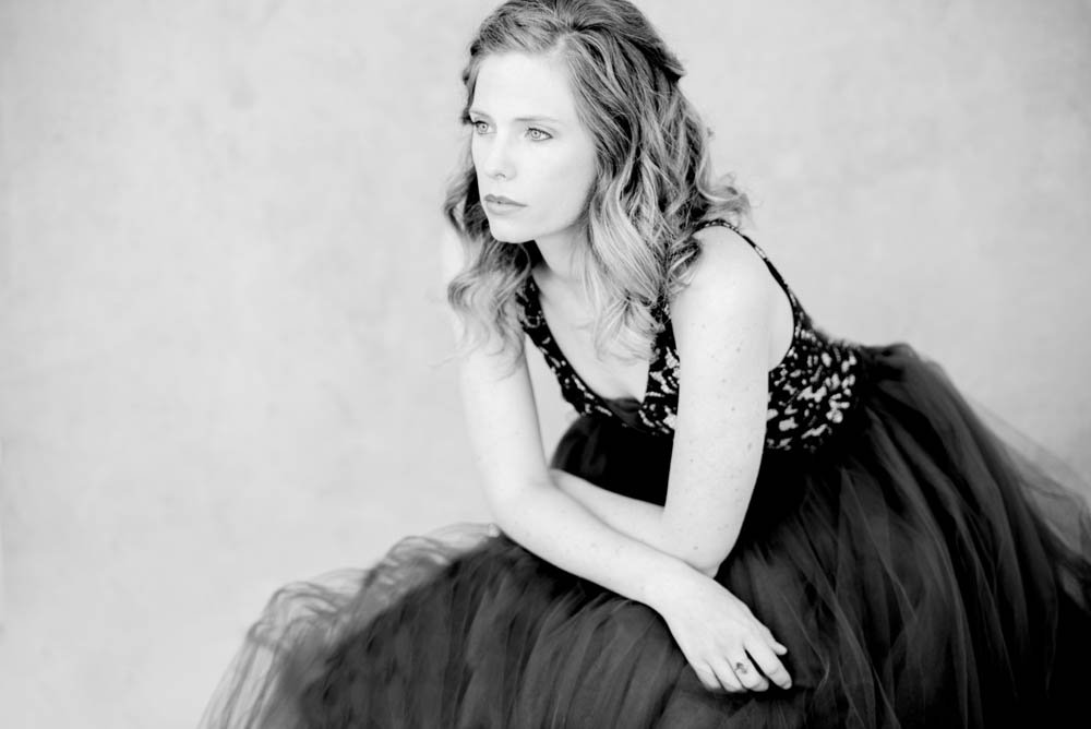A woman with wavy hair in a black floral top and tulle skirt, sitting and looking to the side in a black and white photo.