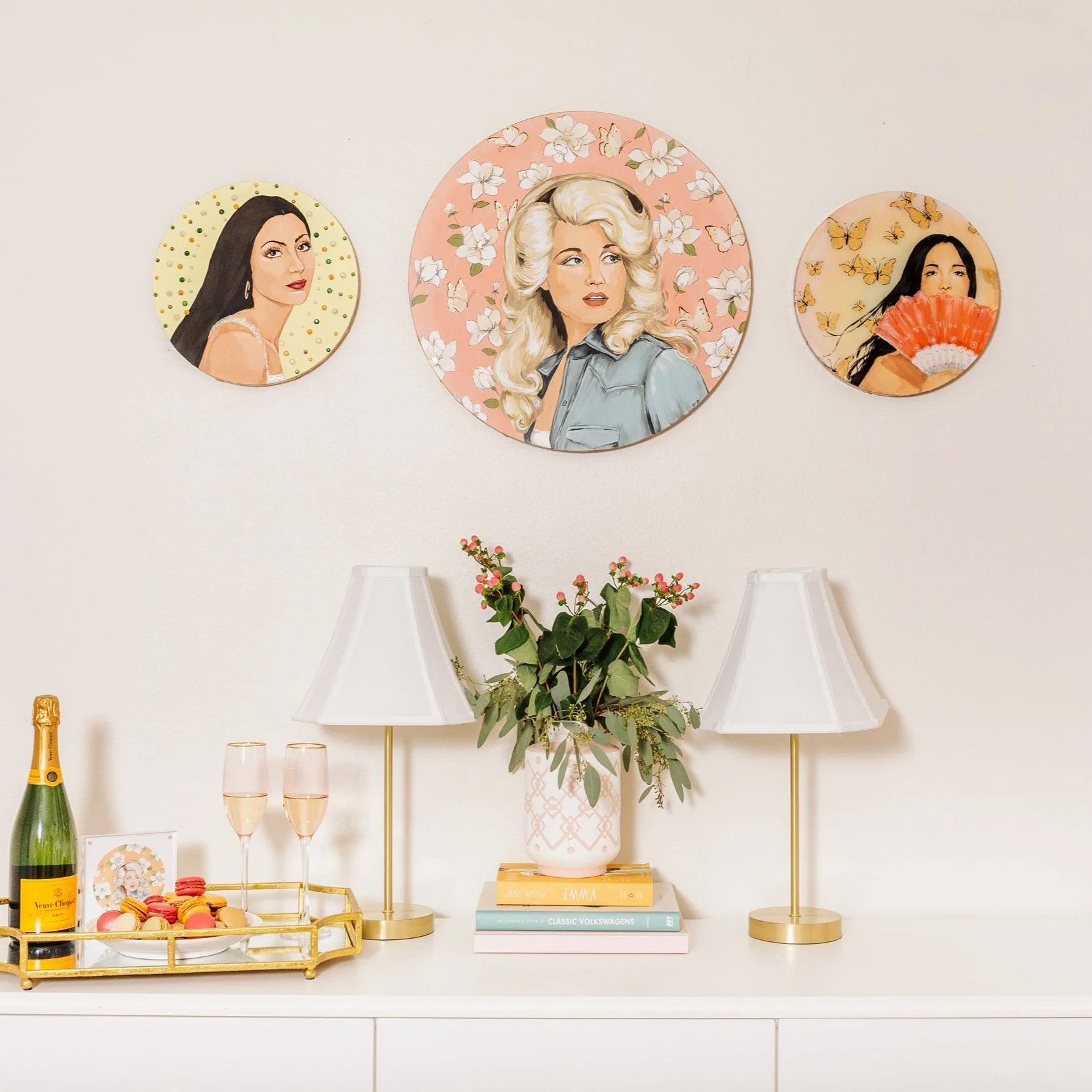 painted celebrity portraits of dolly parton, cher, and kacey musgraves displayed in a room. 