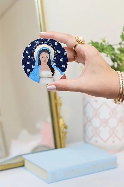 a 3 inch sticker of the virgin mary on a dark navy background surrounded by white flowers and a gold halo.