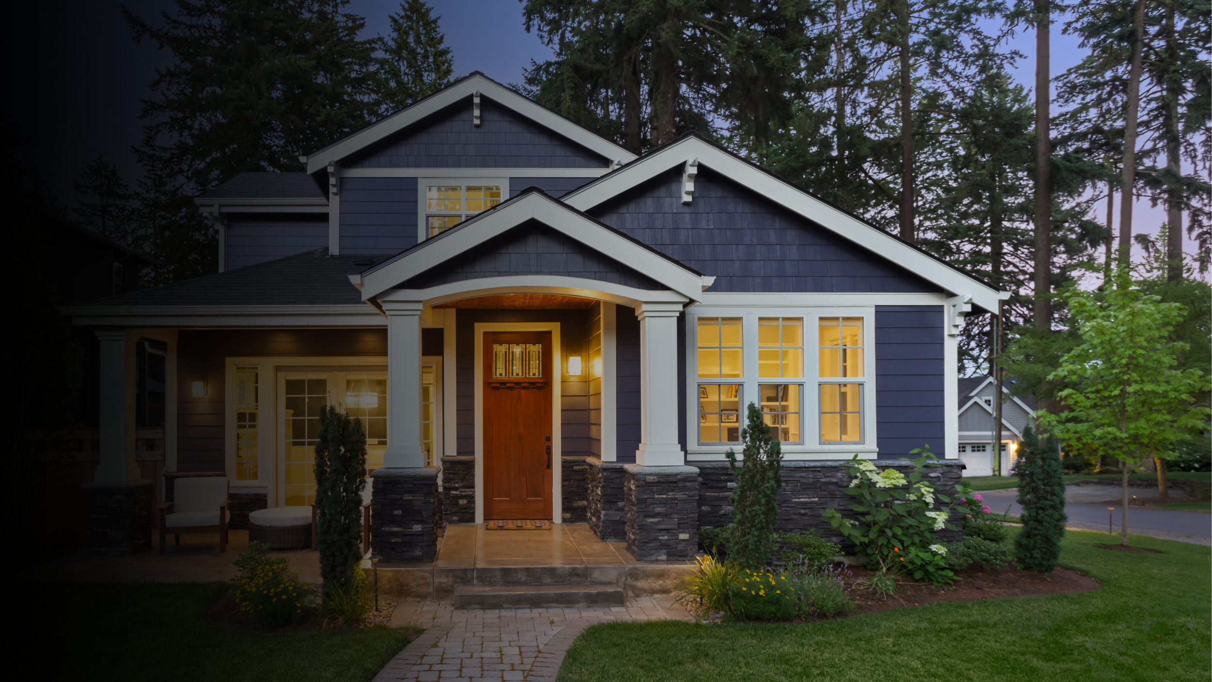 A modern two-story house with blue siding, white trim, and stone accents at the base. The front porch is lit with warm lighting, and the door is wooden. The house has multiple large windows with yellow lighting inside, and a well-maintained front yard with green grass, bushes, and small trees. Tall trees are in the background, and it appears to be dusk or early evening.