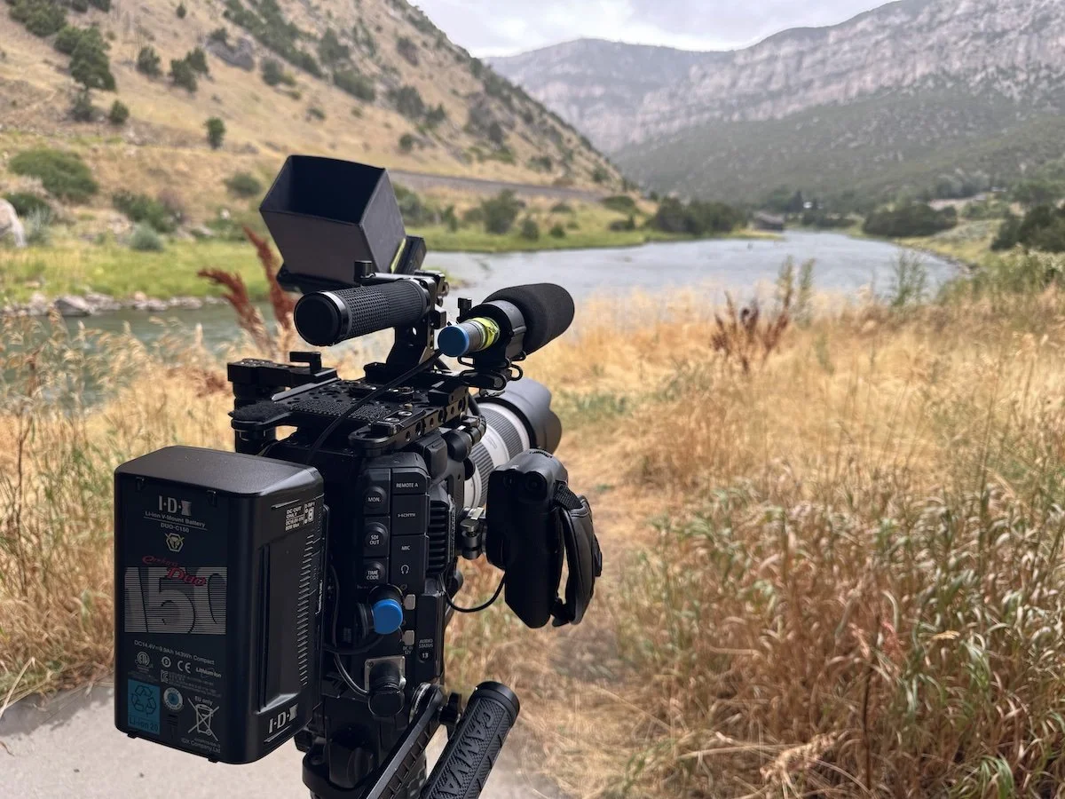 A professional video camera points towards fly fishermen in the distance. Photo taken in the Wind River Canyon in Wyoming.