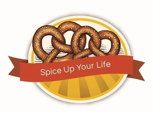A logo featuring a pretzel with a red banner that says "Spice Up Your Life" over a yellow and white circular background.