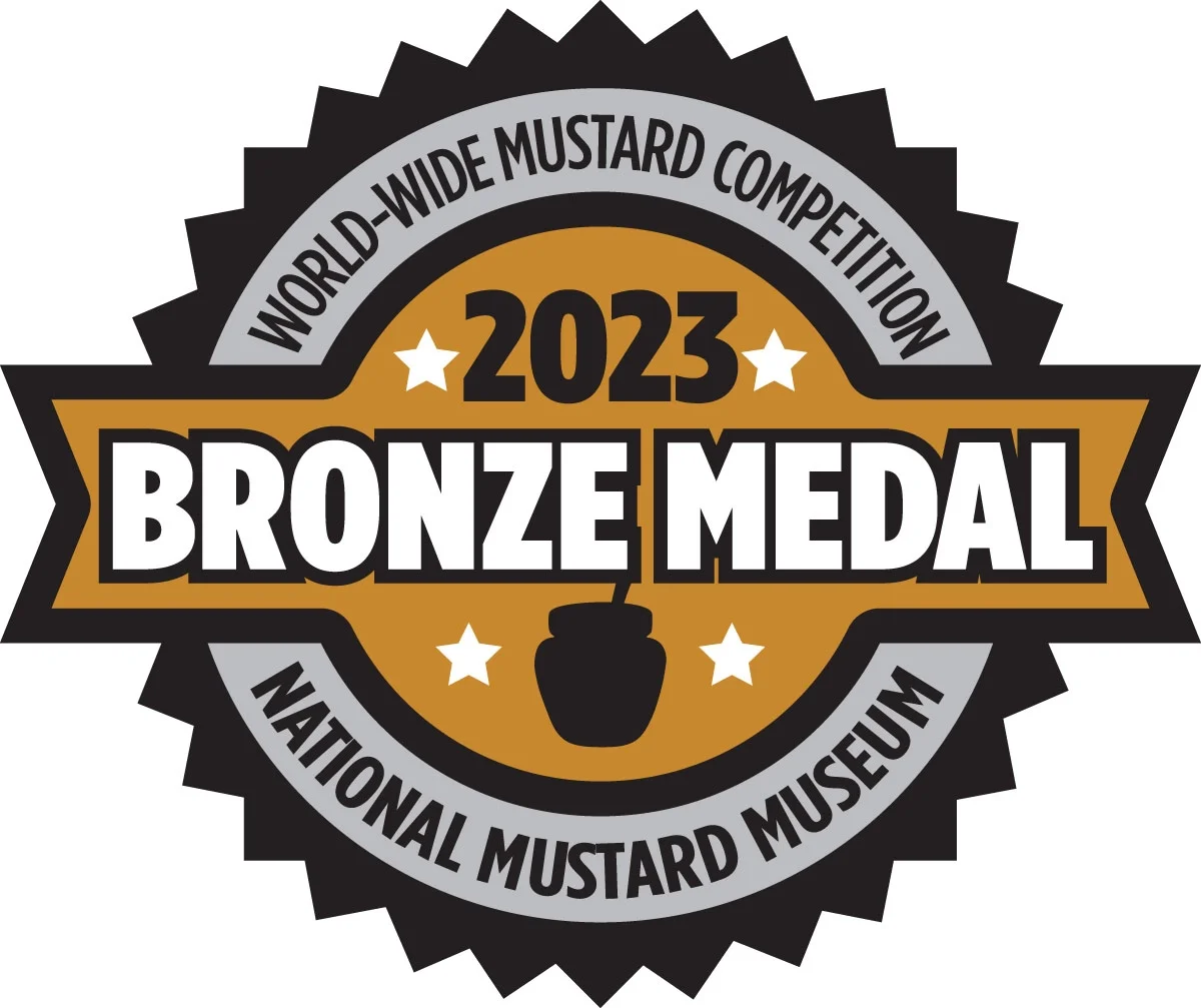 Bronze medal badge from the 2023 World-Wide Mustard Competition at the National Mustard Museum, featuring a black and gold design with a mustard jar icon and star accents won by Suzie's Organic Yellow Mustard.