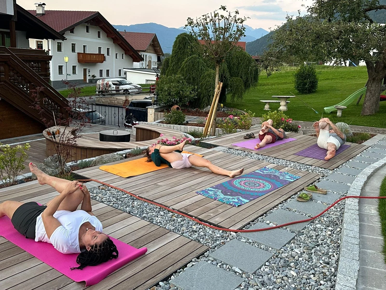 Five people practicing yoga outdoors on mats on a wooden patio in a scenic, mountainous area during the late afternoon or early evening.