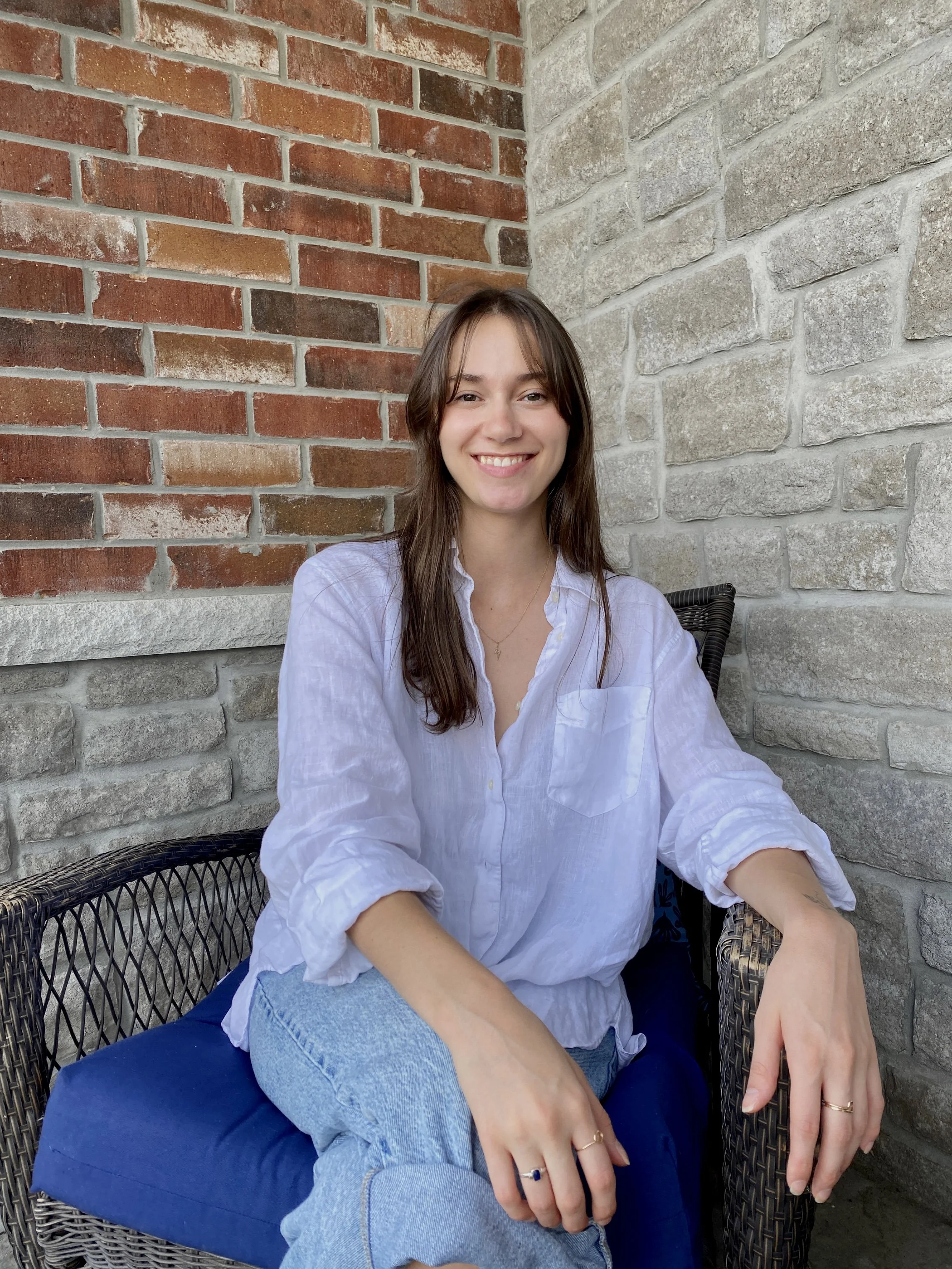 A young woman with long brown hair, wearing a white blouse and blue jeans, sitting on a chair with a blue cushion, smiling at the camera against a brick and stone wall background.