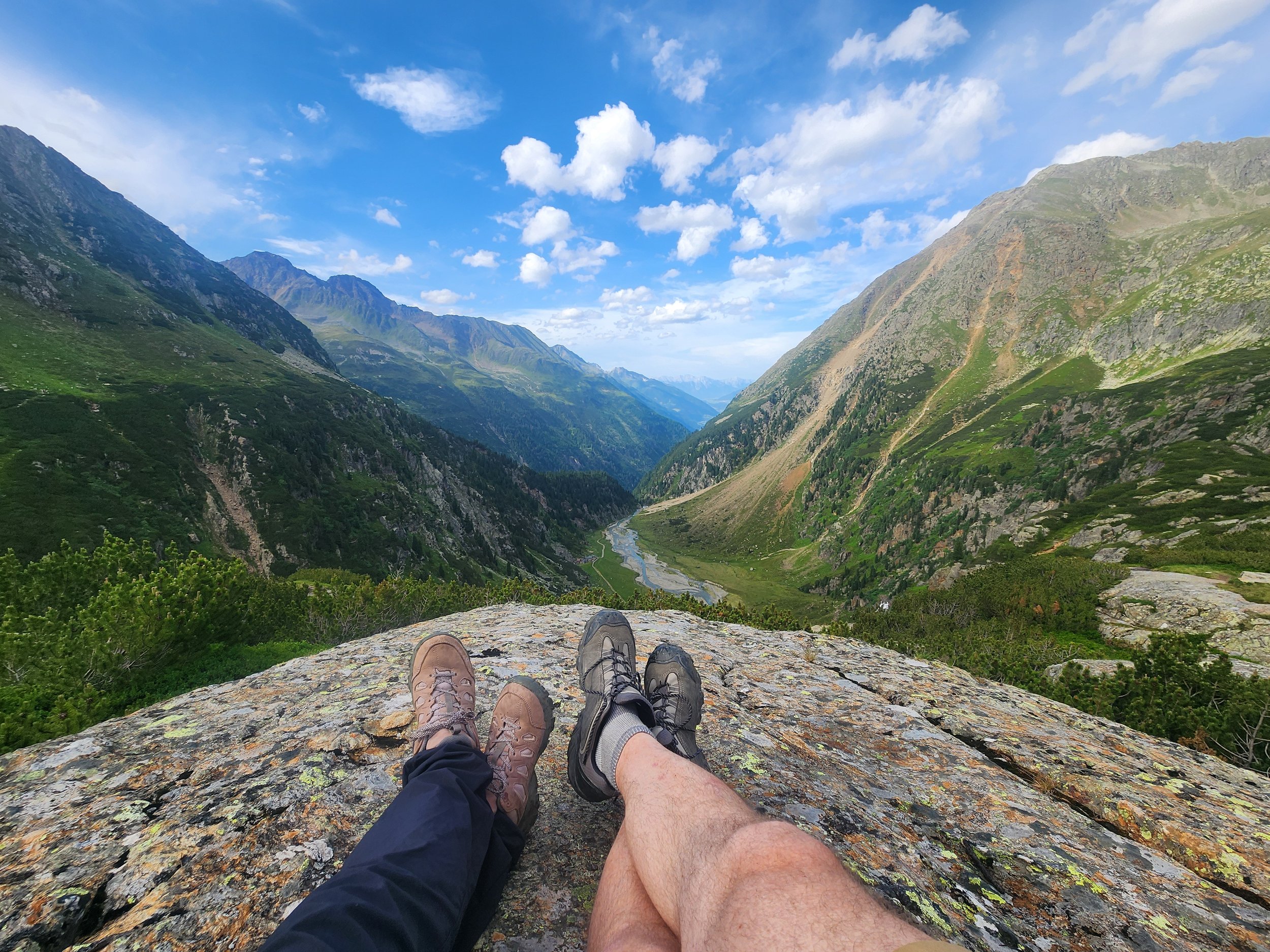 Two hikers sitting on a rocky ledge overlooking a mountain valley with green slopes, a winding river, and a blue sky with scattered clouds.