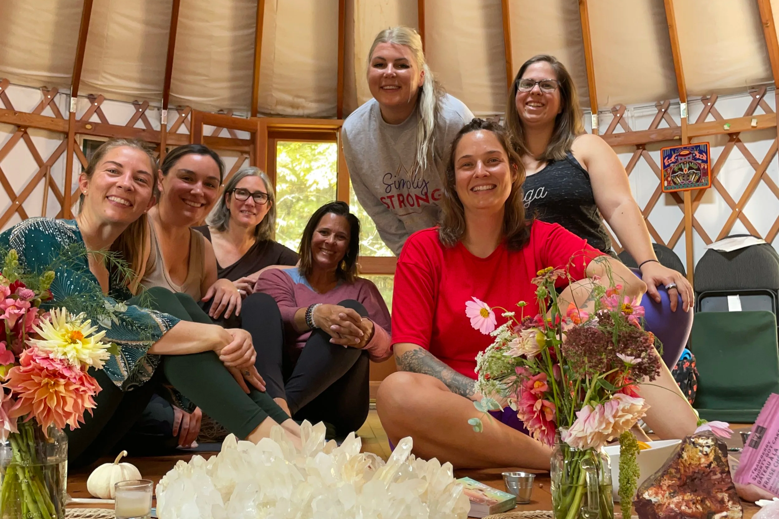Group of eight women smiling and sitting around a table with flowers, candles, and decorative items, inside a wooden dome-shaped structure with a window showing trees outside.