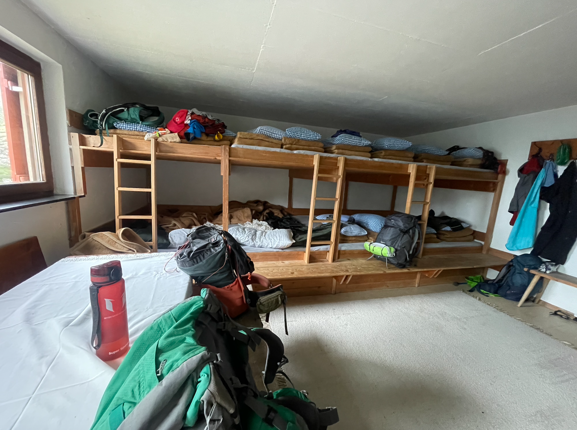 The room contains a bunk bed with multiple sleeping bags and pillows, backpacks, and duffel bags. There is a table with a red water bottle and hiking gear, and a window on the left. Clothes hang on hooks on the right wall, and the floor is partially 