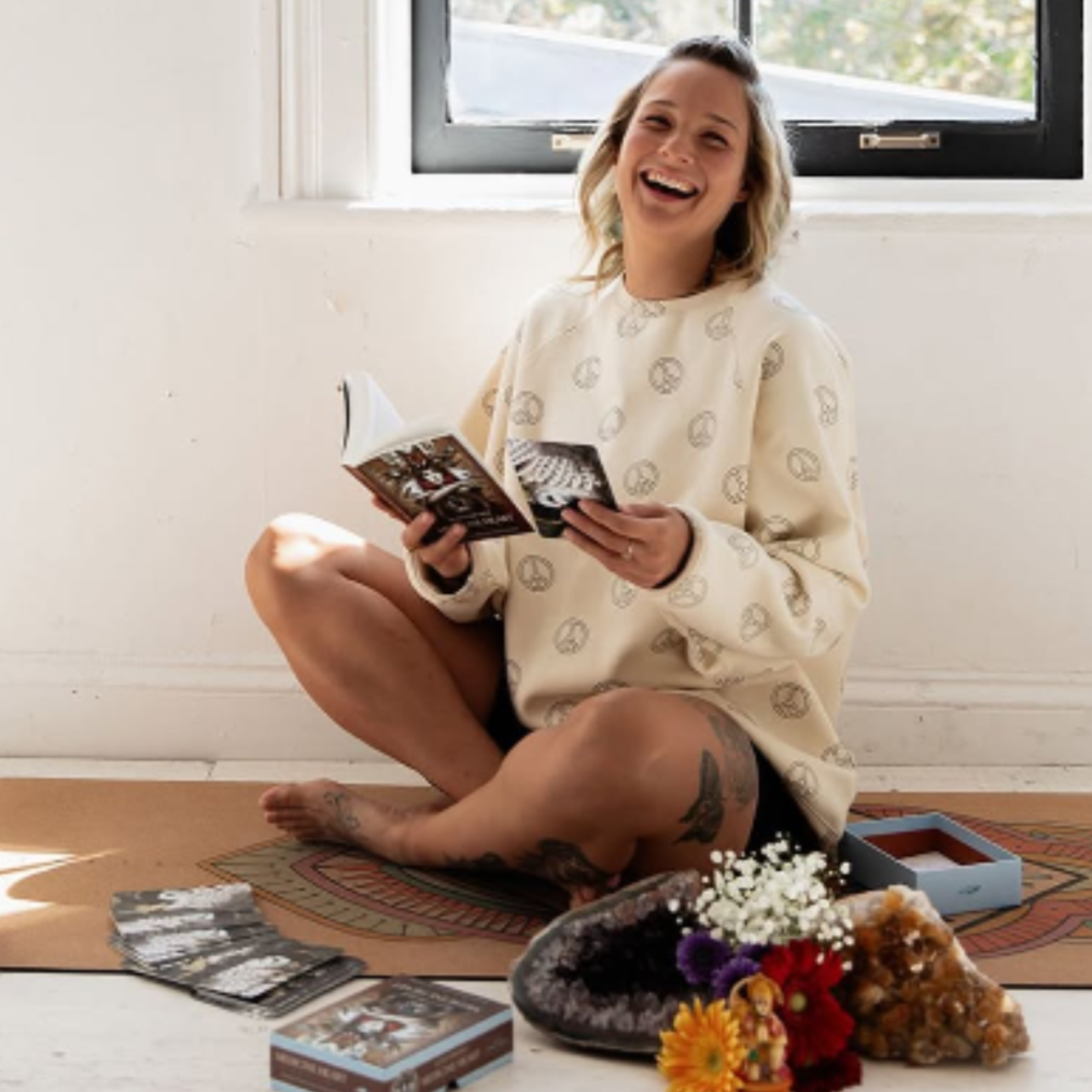 A woman with tattoos on her legs, wearing a cream sweater, is sitting cross-legged on the floor next to a window. She is smiling and reading a card. There are tarot cards, a box, flowers, and a geode on the floor in front of her.