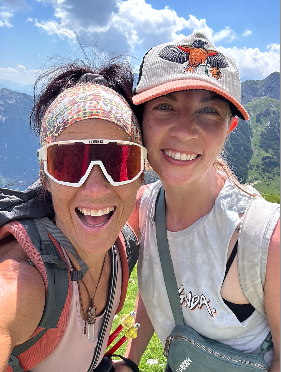 Two women in outdoor mountain setting, smiling, wearing hiking gear and hats, with lush green mountains in the background.