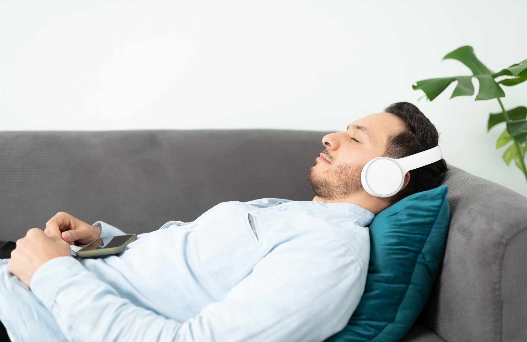 A man with closed eyes, relaxed and wearing white headphones, lying on a gray sofa with a blue pillow, holding a smartphone.
