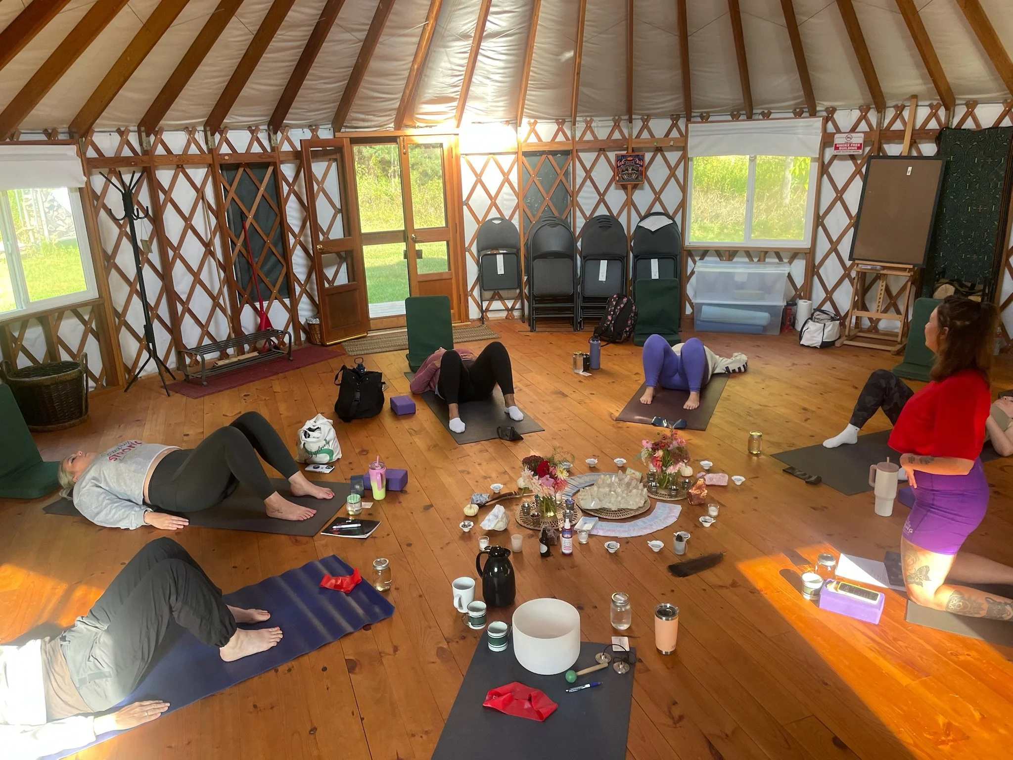 People participating in a yoga class inside a wooden yurt, lying on yoga mats on a wooden floor, with meditation and altar items in the center, and windows showing greenery outside.