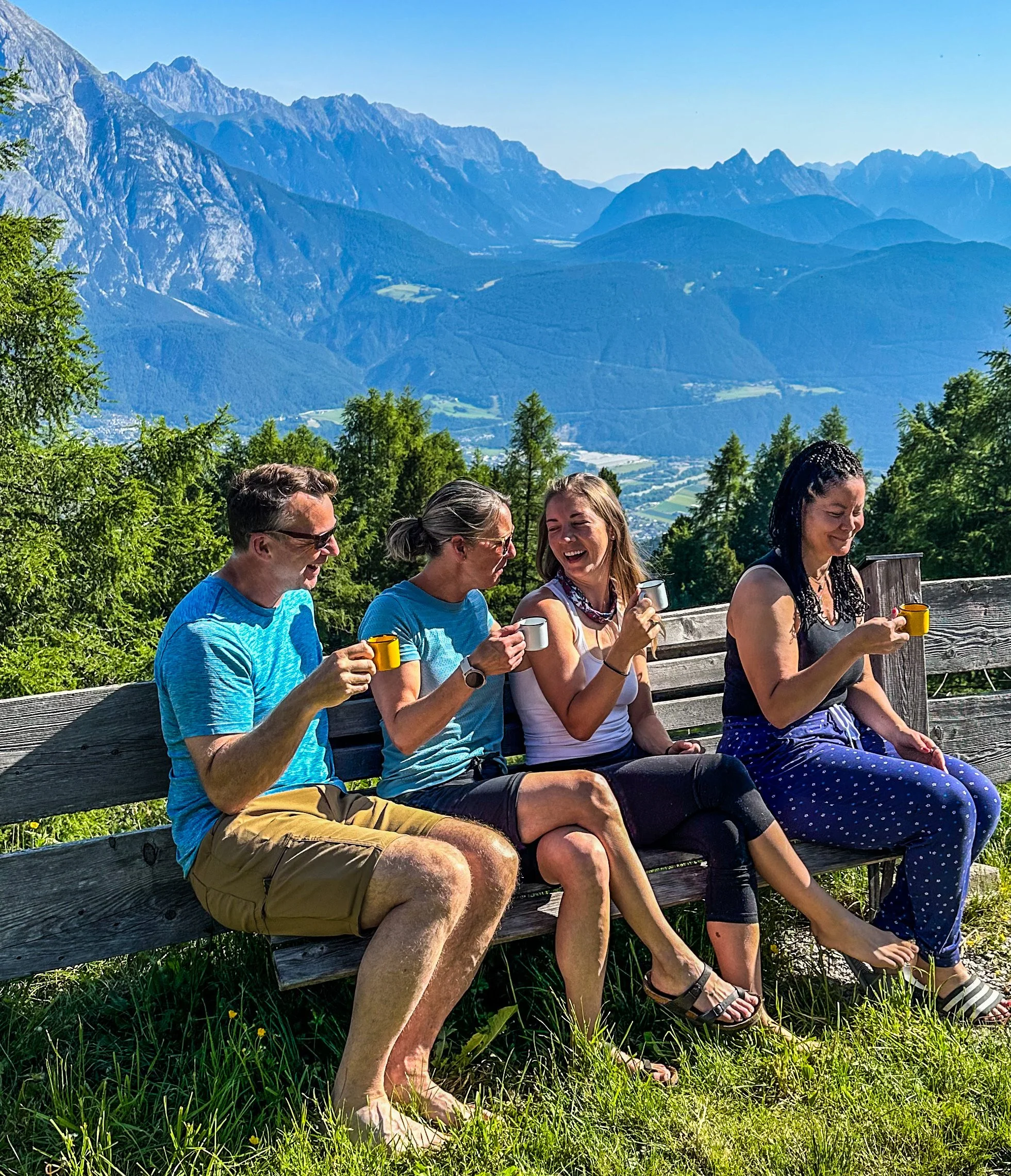Four people sitting on a wooden bench outdoors, enjoying coffee with mountain scenery in the background.