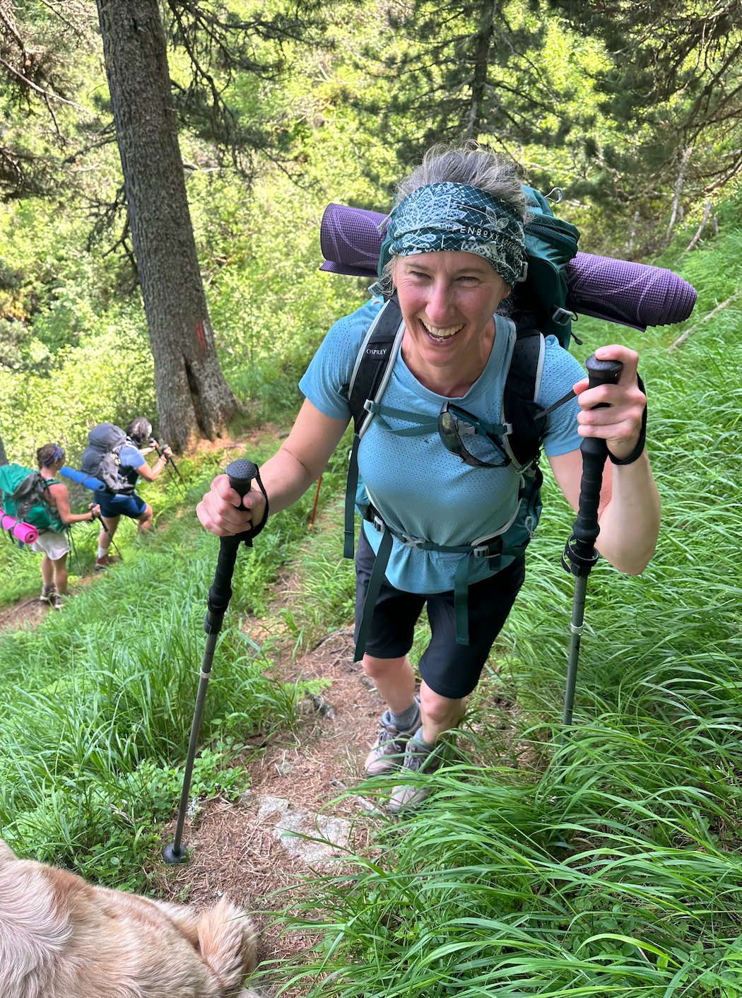 A woman hiking on a forest trail with a pack and hiking poles, smiling, with other hikers in the background.