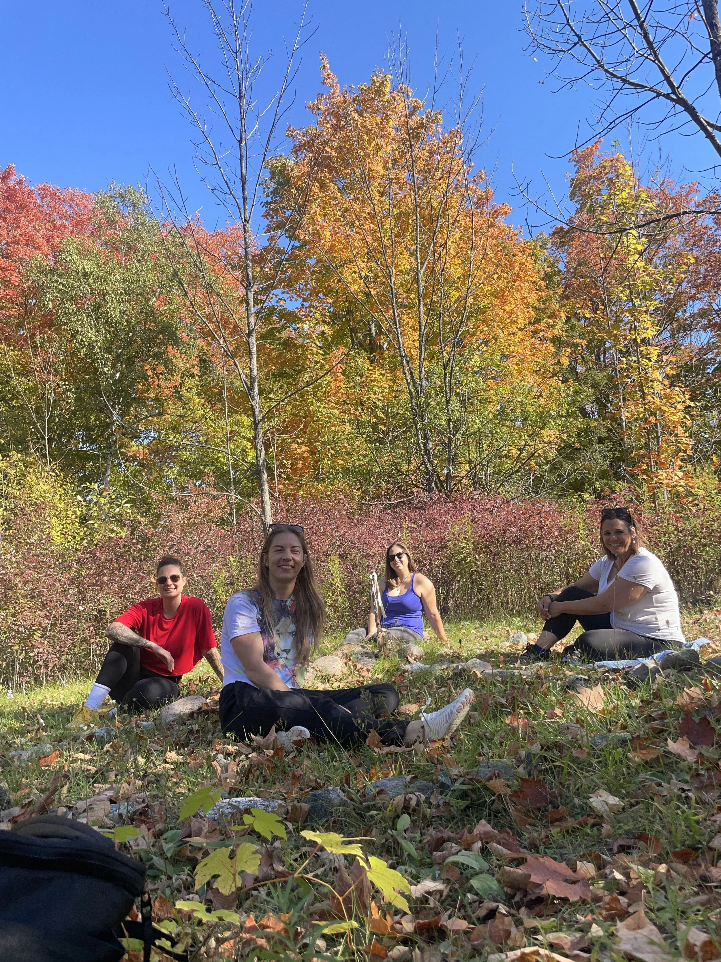 Four women sitting on the ground in a forest during fall with colorful autumn leaves, blue sky, and trees in the background.