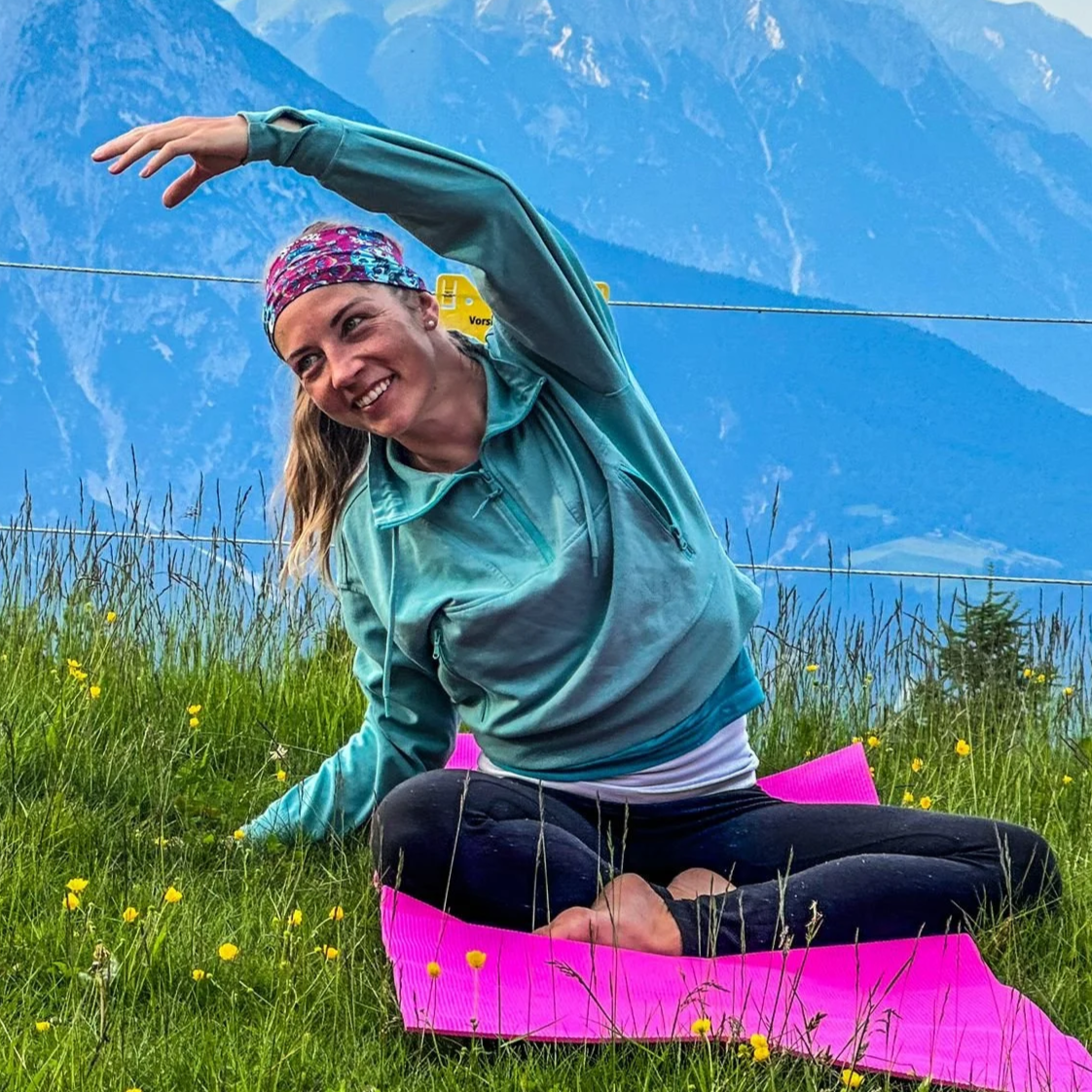 A woman practicing yoga outdoors on a pink yoga mat on a grassy field with yellow flowers, mountains in the background, smiling and twisting in a seated side stretch pose.
